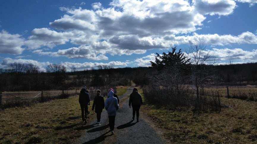 Under a brilliant blue cloud streaked sky, five teens walking away from the camera on a path between two fields